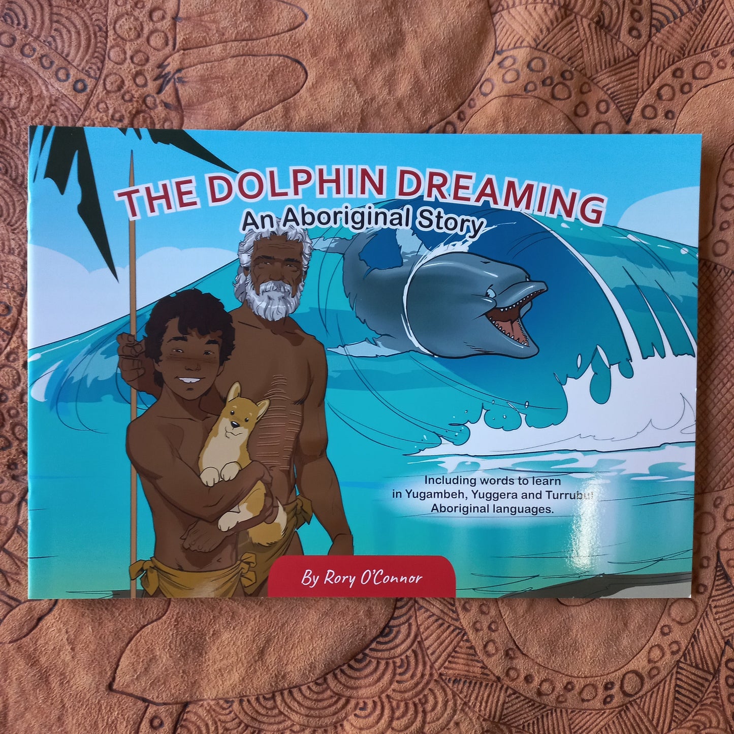 A children's book titled "The Dolphin Dreaming: An Aboriginal Story" by Rory O'Connor lies flat on a brown patterned fabric surface. The cover features colorful artwork of a dolphin, a bearded man, a woman, and children, with a red vertical banner reading "By Rory O'Connor.