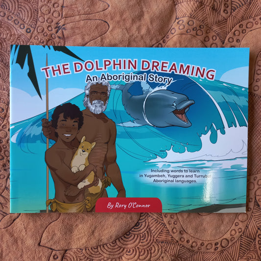 A children's book titled "The Dolphin Dreaming: An Aboriginal Story" by Rory O'Connor lies flat on a brown patterned fabric surface. The cover features colorful artwork of a dolphin, a bearded man, a woman, and children, with a red vertical banner reading "By Rory O'Connor.